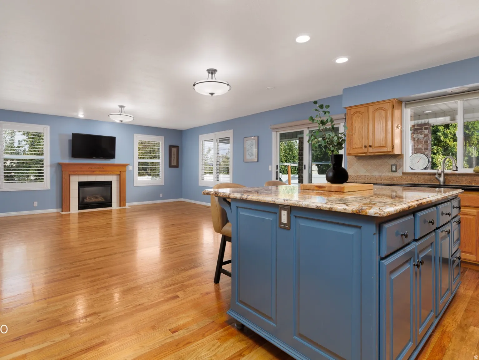Hardwood flooring, kitchen island with stone countertop, built-in fireplace with wood mantel, multiple windows with white shutters, and sliding glass doors