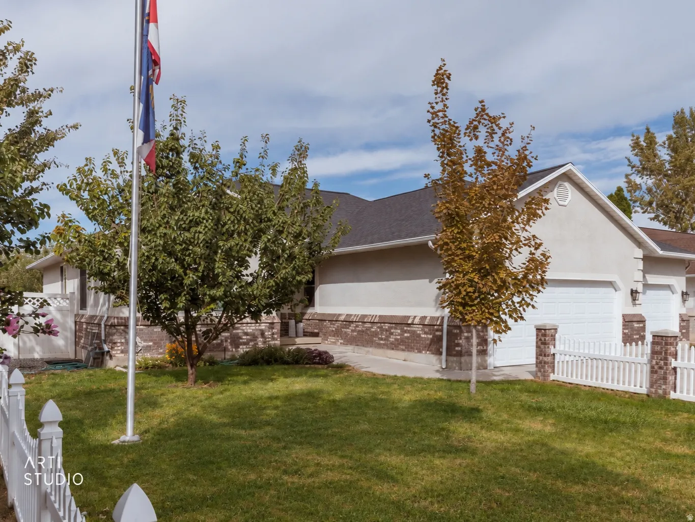 Stucco exterior with brick detailing, white picket fence, attached garage, and mature landscaping