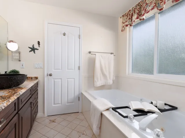 Expansive soaking tub with window, wood-finish vanity with stone countertop, and vessel sink with wall-mounted faucet