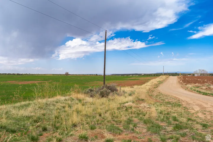 Expansive rural landscape featuring a dirt road, open fields, scattered low-lying vegetation, and utility poles