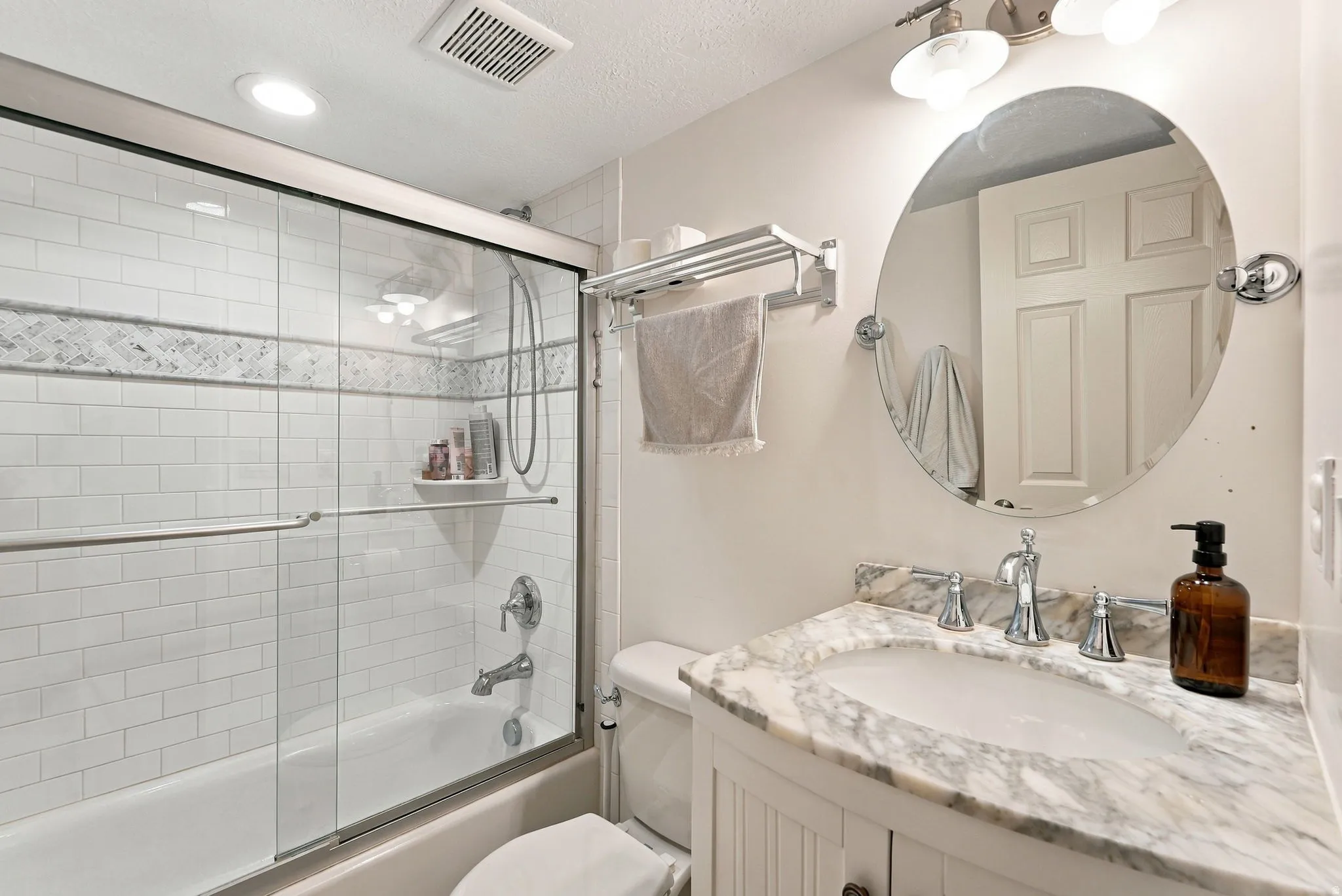 Bathroom featuring a vanity with a stone countertop, a circular mirror, a tub with subway tile surround, a decorative tile accent band, and a glass shower enclosure