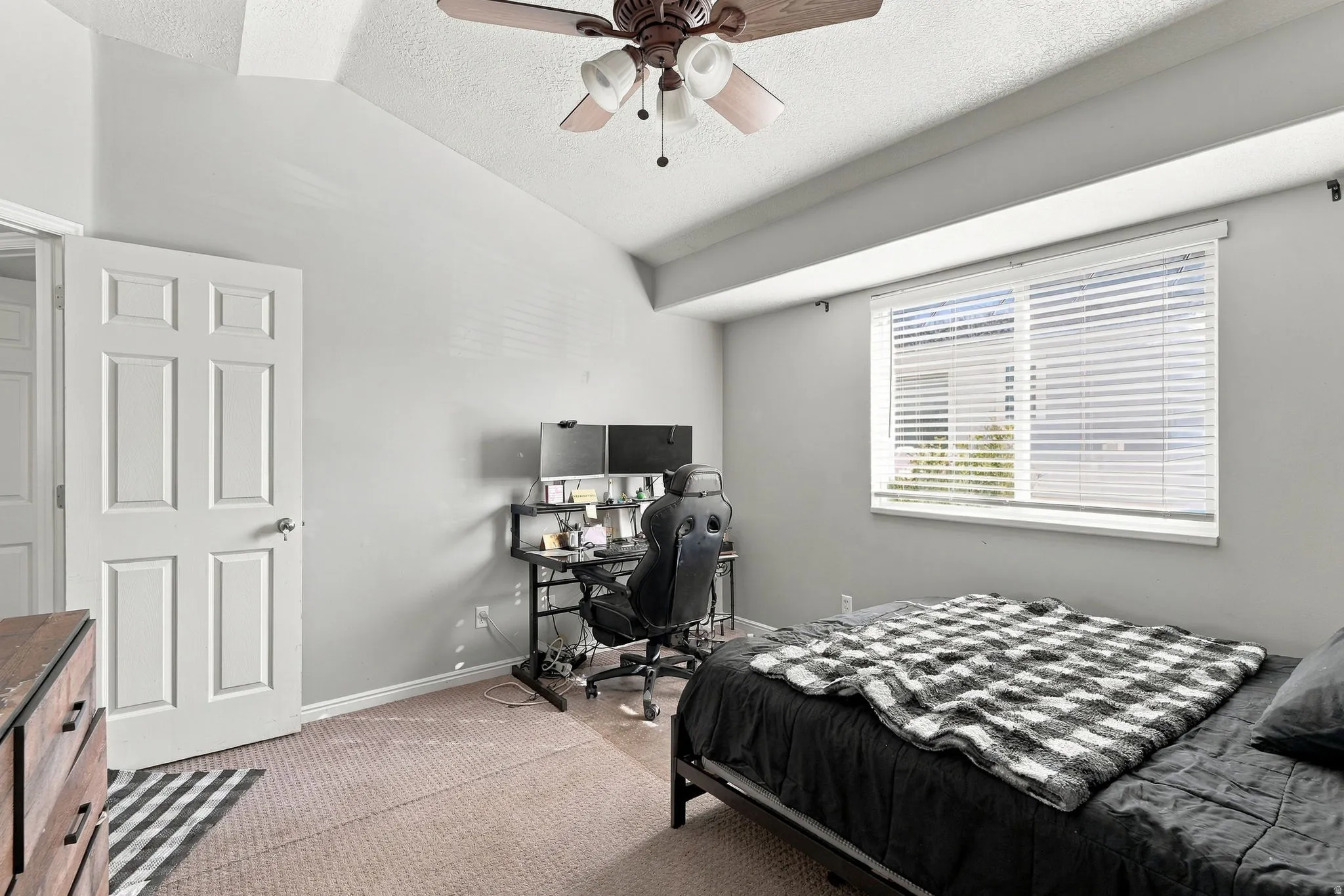 Vaulted ceiling, ceiling fan with integrated lighting, paneled interior door, carpet flooring, and a large window with blinds