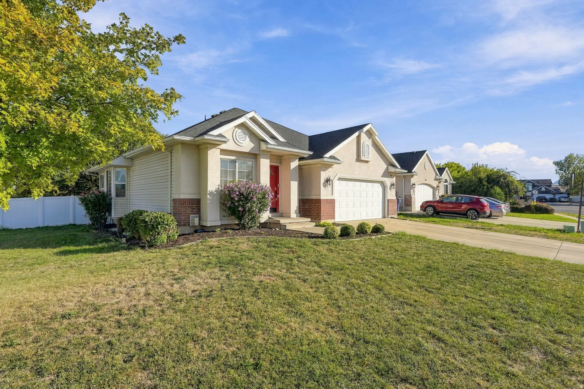 Stucco and brick facade with an attached two-car garage, mature landscaping, a white privacy fence, and a red entry door