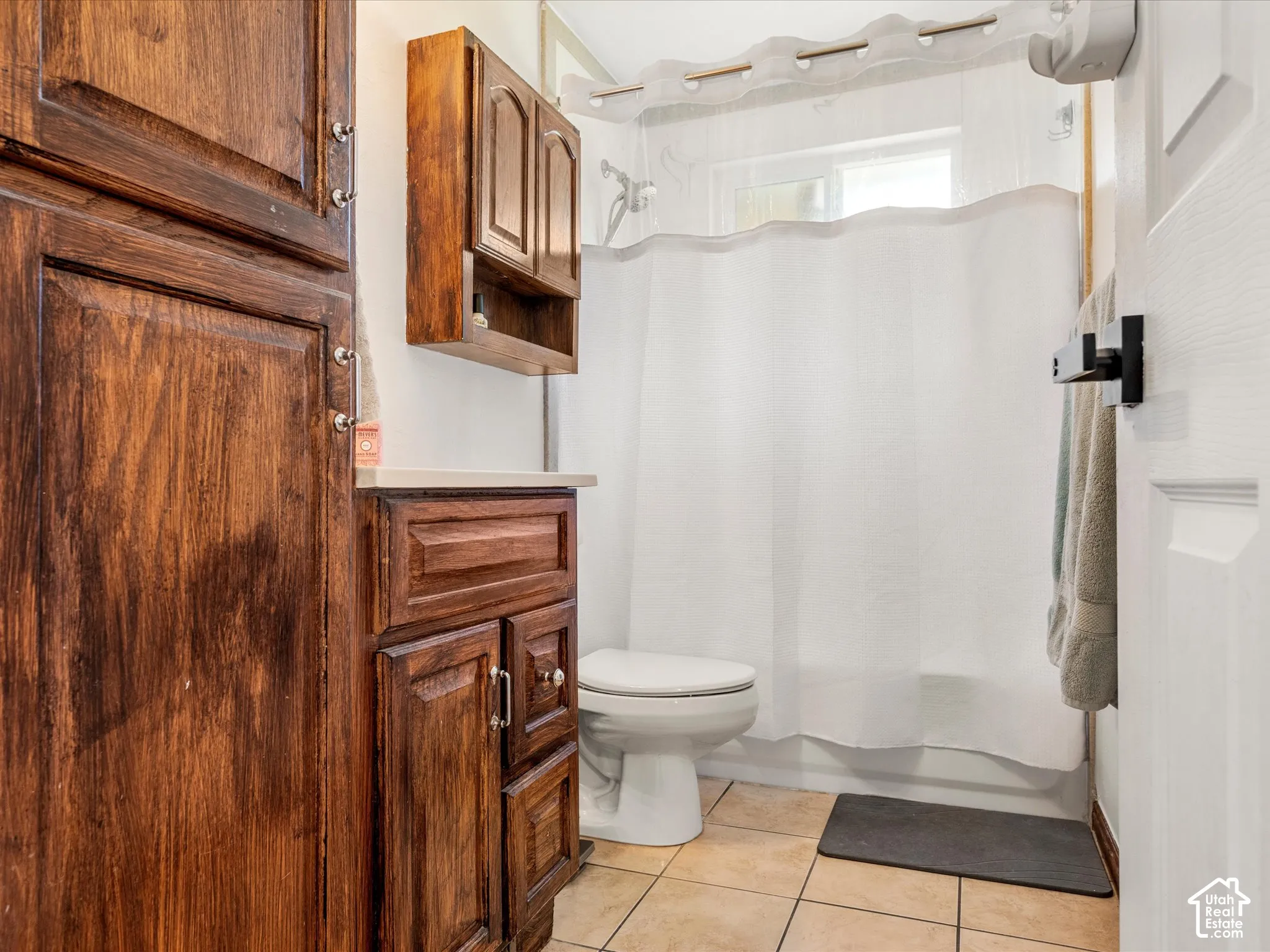 Wood-finish cabinetry, light-toned countertop, white toilet, tiled flooring, and a window for natural light