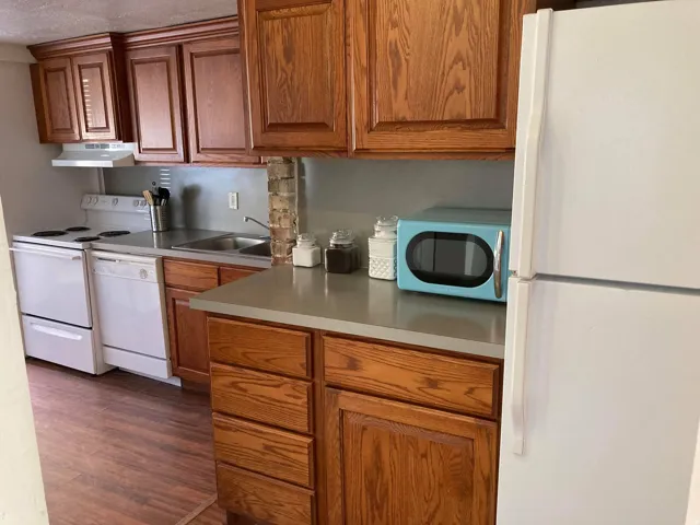 Kitchen featuring wood-finish cabinetry, solid surface countertops, a stainless steel sink, white appliances, and wood-finish flooring