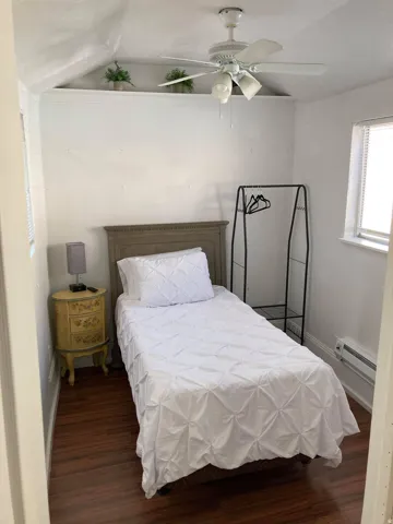 Wood-finish flooring, white ceiling fan with integrated lighting, window with blinds, baseboard heater, and a built-in display shelf