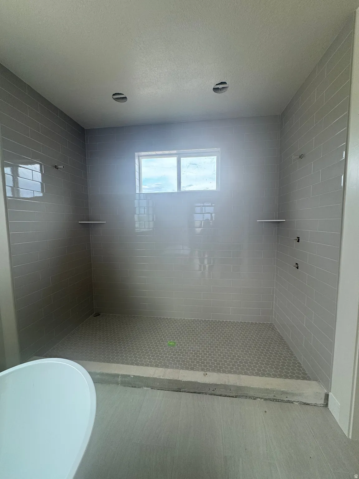 Expansive walk-in shower featuring light grey subway tile walls, a grey hexagon tile floor, and a window for natural light