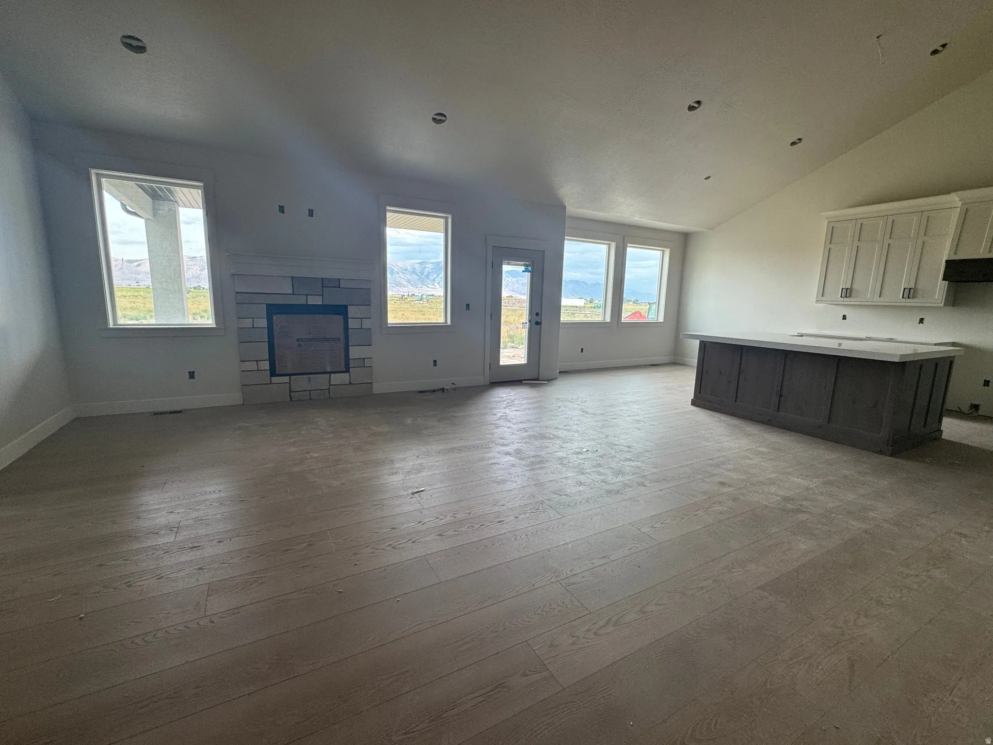 Vaulted ceiling, stone fireplace with mantel, white shaker cabinetry, wood-finish flooring, and a large island with a light-colored countertop