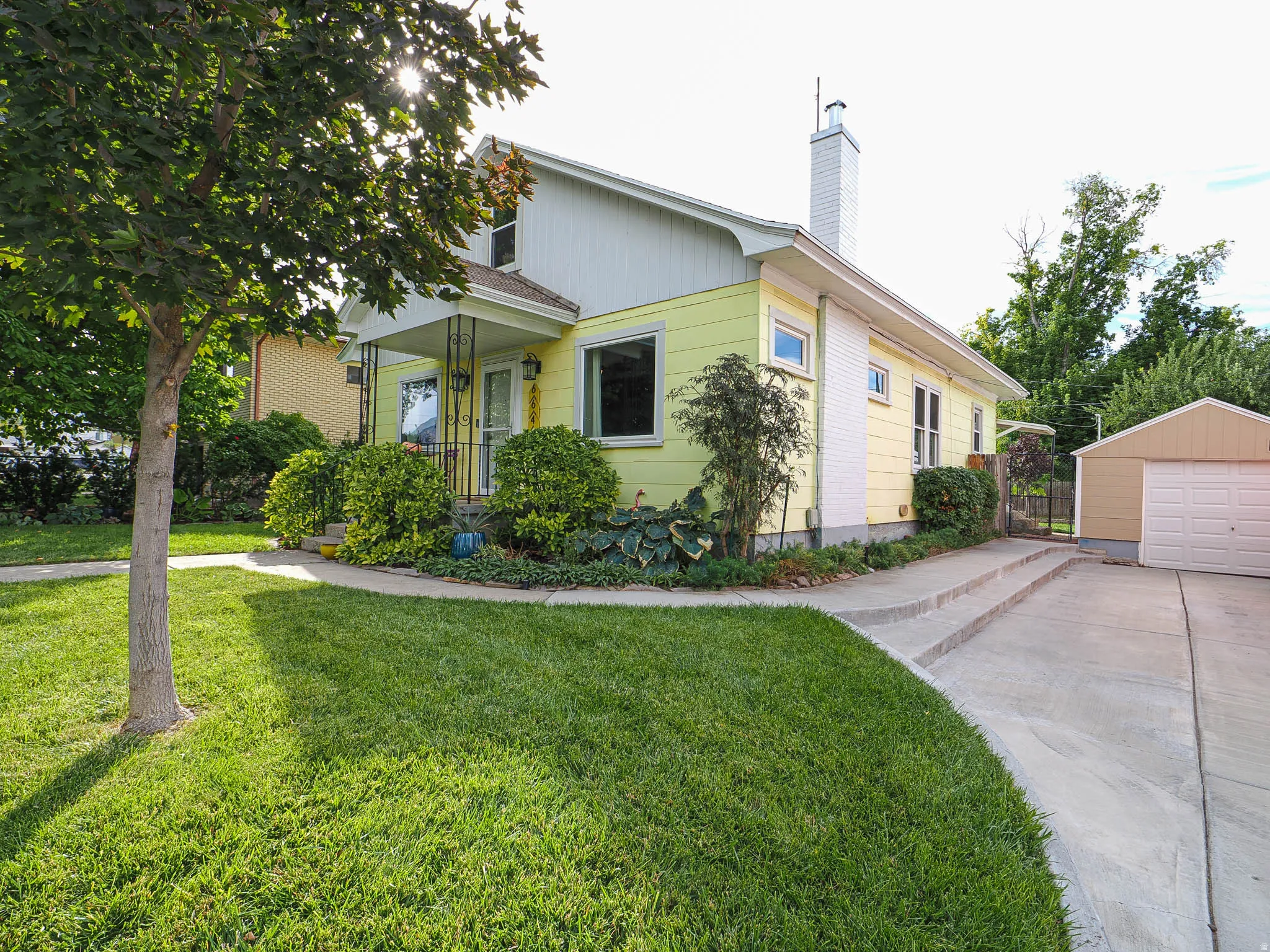 Exterior featuring light yellow siding, a covered porch with decorative railing, mature landscaping, a concrete driveway, and a detached garage
