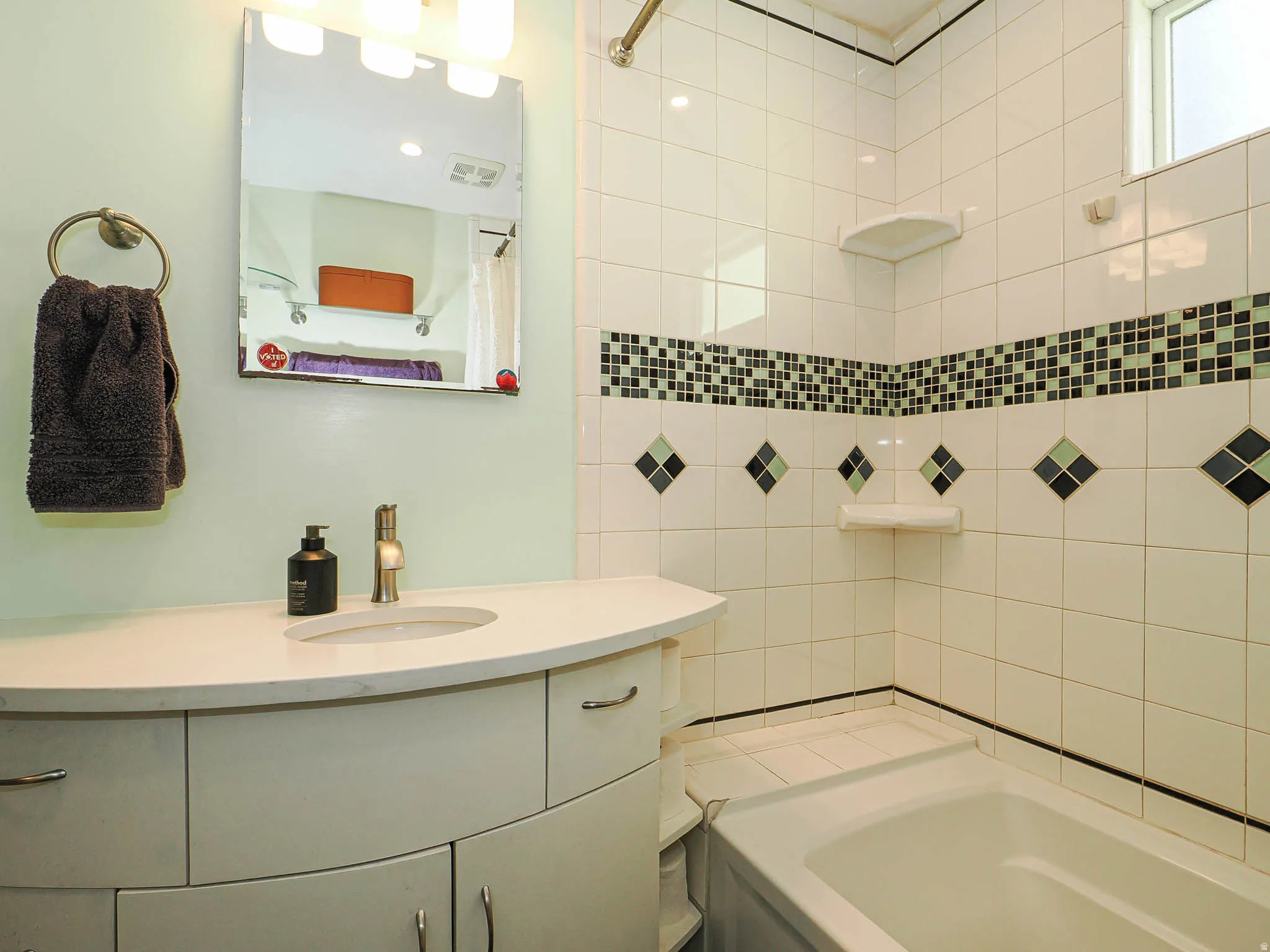 Vanity with a curved countertop and integrated sink, white tiled walls with decorative accents, an overhead light fixture, and a built-in bathtub