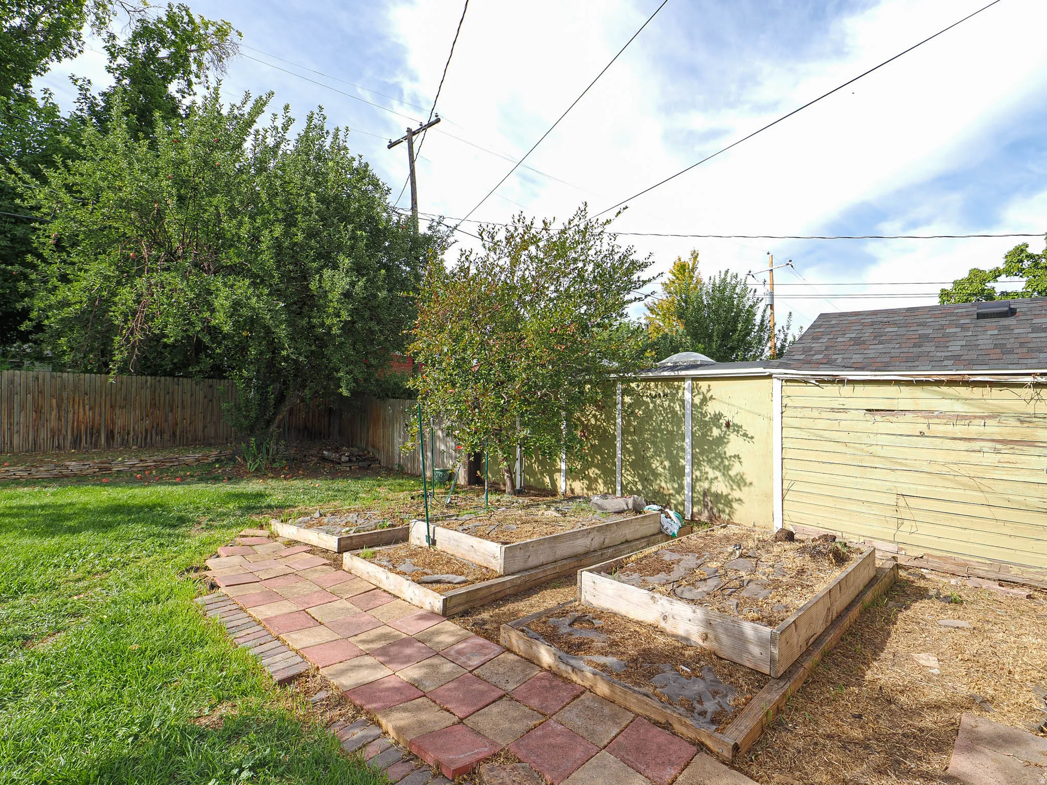 Expansive outdoor space featuring a paved pathway, multiple raised garden beds, mature trees, and a wood privacy fence