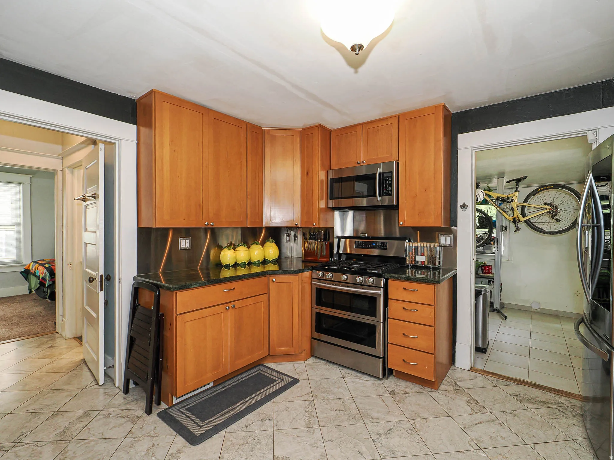 Wood cabinetry with under-cabinet lighting, stainless steel appliances, dark stone countertops, tiled flooring, and a flush mount ceiling light