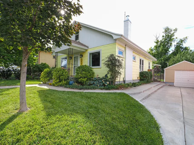 Exterior featuring light yellow siding, a covered porch with decorative railing, mature landscaping, a concrete driveway, and a detached garage