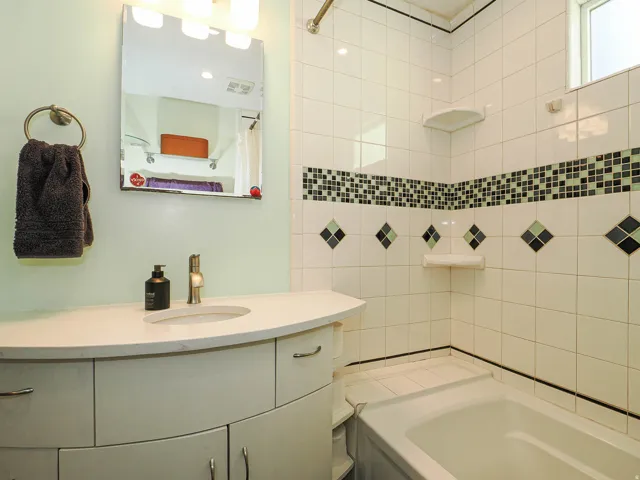 Vanity with a curved countertop and integrated sink, white tiled walls with decorative accents, an overhead light fixture, and a built-in bathtub