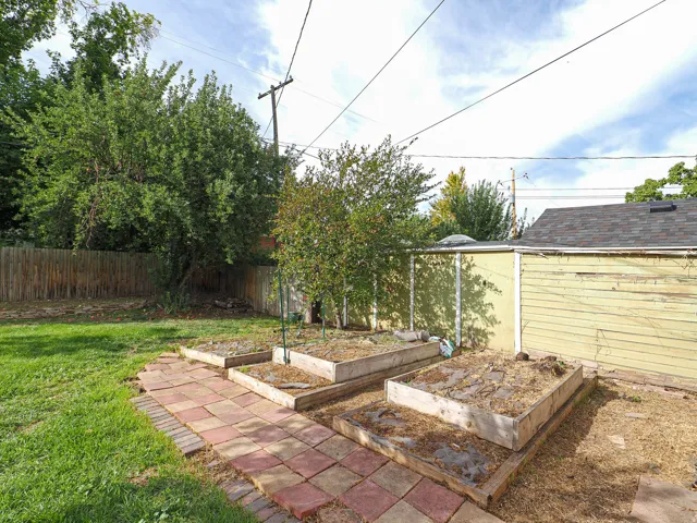 Expansive outdoor space featuring a paved pathway, multiple raised garden beds, mature trees, and a wood privacy fence