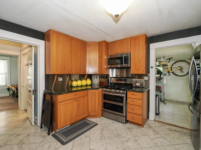 Wood cabinetry with under-cabinet lighting, stainless steel appliances, dark stone countertops, tiled flooring, and a flush mount ceiling light