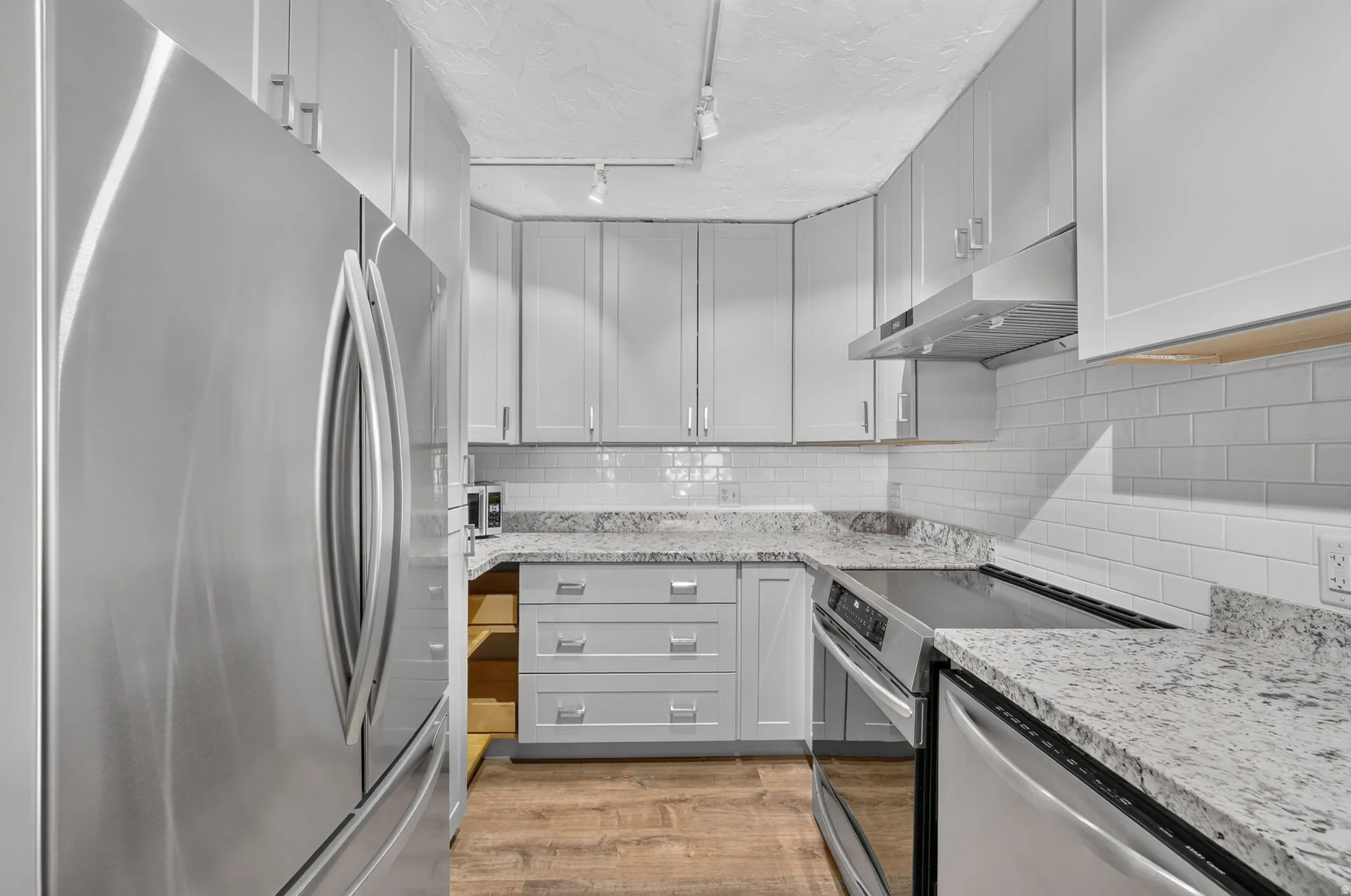 Stainless steel appliances, wood-finish flooring, white subway tile backsplash, white shaker-style cabinetry, and light-toned countertops