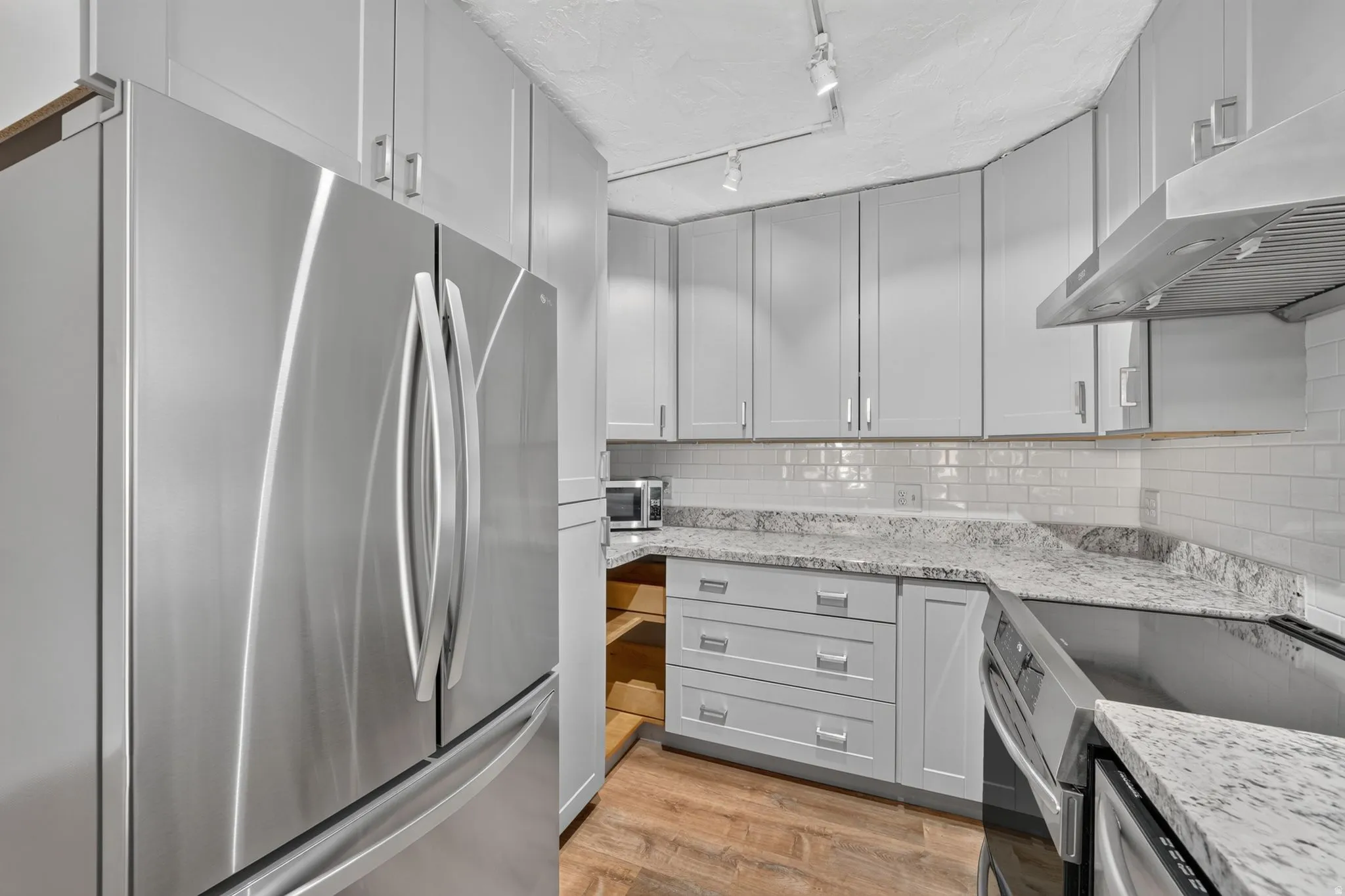 Stainless steel refrigerator, light gray cabinetry with brushed nickel hardware, white subway tile backsplash, speckled countertops, and wood-finish flooring