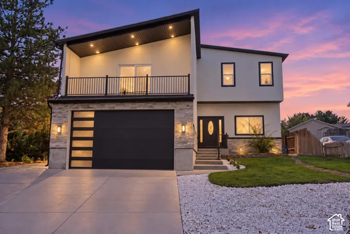 Modern multi-level exterior featuring a slanted roofline, stone veneer accents, second-story balcony with railings, and an oversized garage door with horizontal glass panels