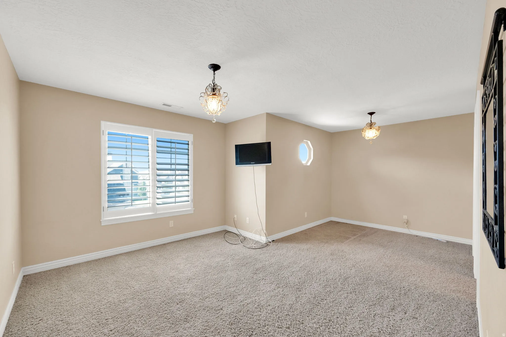 Carpeted room with a large window featuring white plantation shutters, two decorative ceiling light fixtures, and a circular wall cut-out