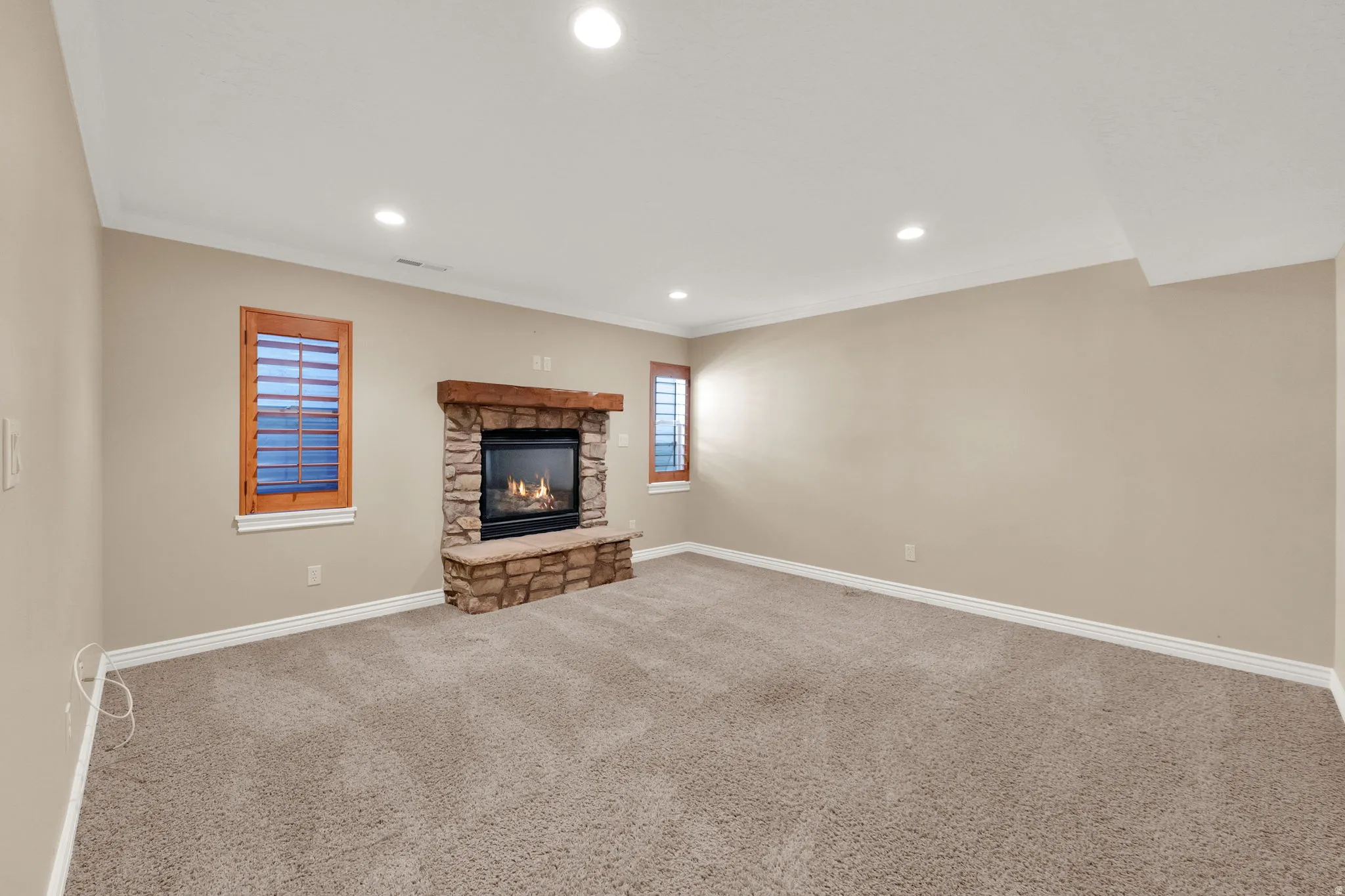 Recessed lighting, stone-clad fireplace with wood mantel, built-in fireplace bench, two windows with wood shutters, and neutral carpeting