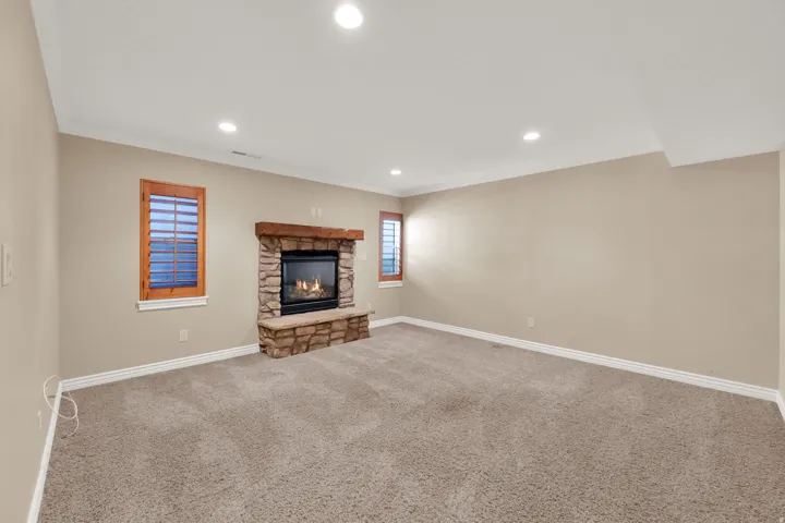 Recessed lighting, stone-clad fireplace with wood mantel, built-in fireplace bench, two windows with wood shutters, and neutral carpeting