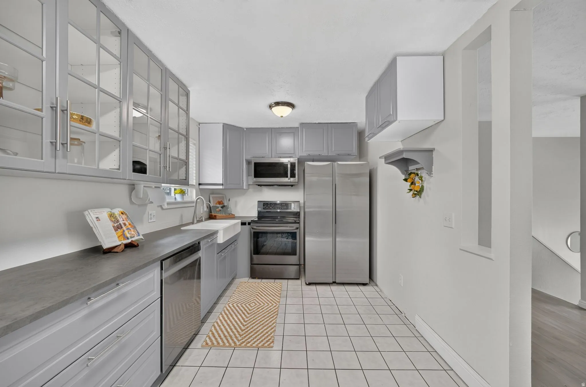 Kitchen featuring gray cabinetry, stainless steel appliances, a white farmhouse sink, light-toned tile flooring, and dark countertops