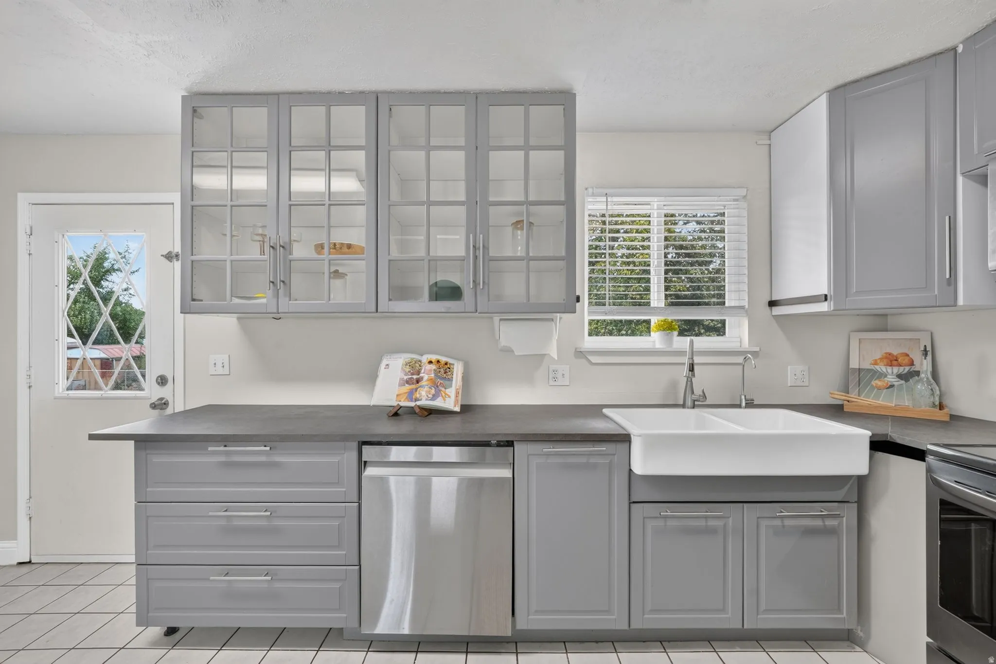 Modern kitchen featuring gray cabinetry with glass-front upper cabinets, a large white farmhouse-style sink with a gooseneck faucet, stainless steel dishwasher, and dark countertops