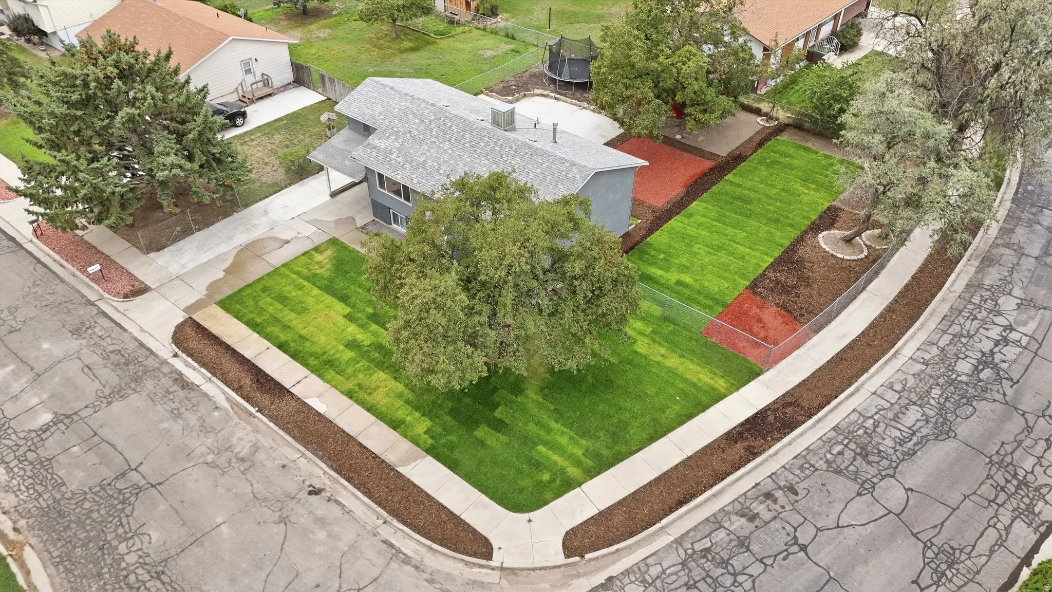 Corner lot property featuring a gray-sided residence with a gray shingle roof, established trees, and a well-maintained lawn with landscaping