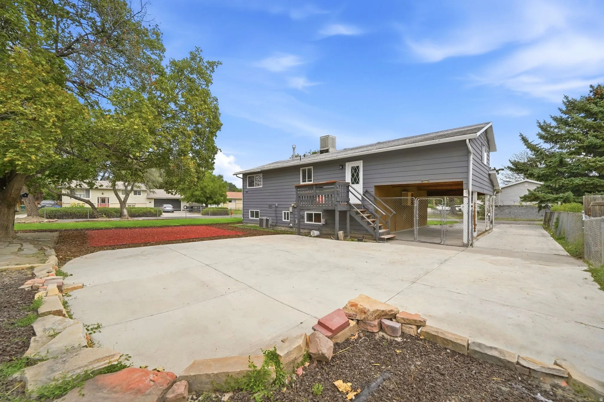 Gray siding exterior with white trim, elevated wood deck with stairs, concrete driveway, and established trees