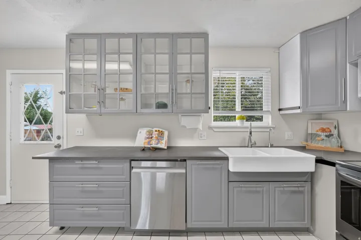 Modern kitchen featuring gray cabinetry with glass-front upper cabinets, a large white farmhouse-style sink with a gooseneck faucet, stainless steel dishwasher, and dark countertops