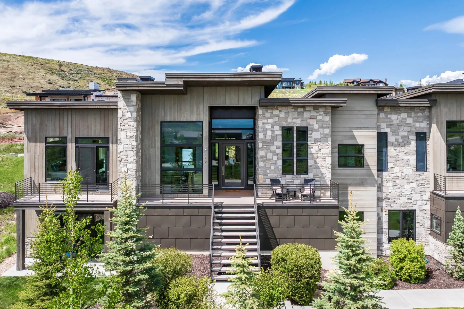 Modern architectural design with stone and wood panel exterior, multiple balconies with metal railings, and a prominent front entrance with full-light glass doors