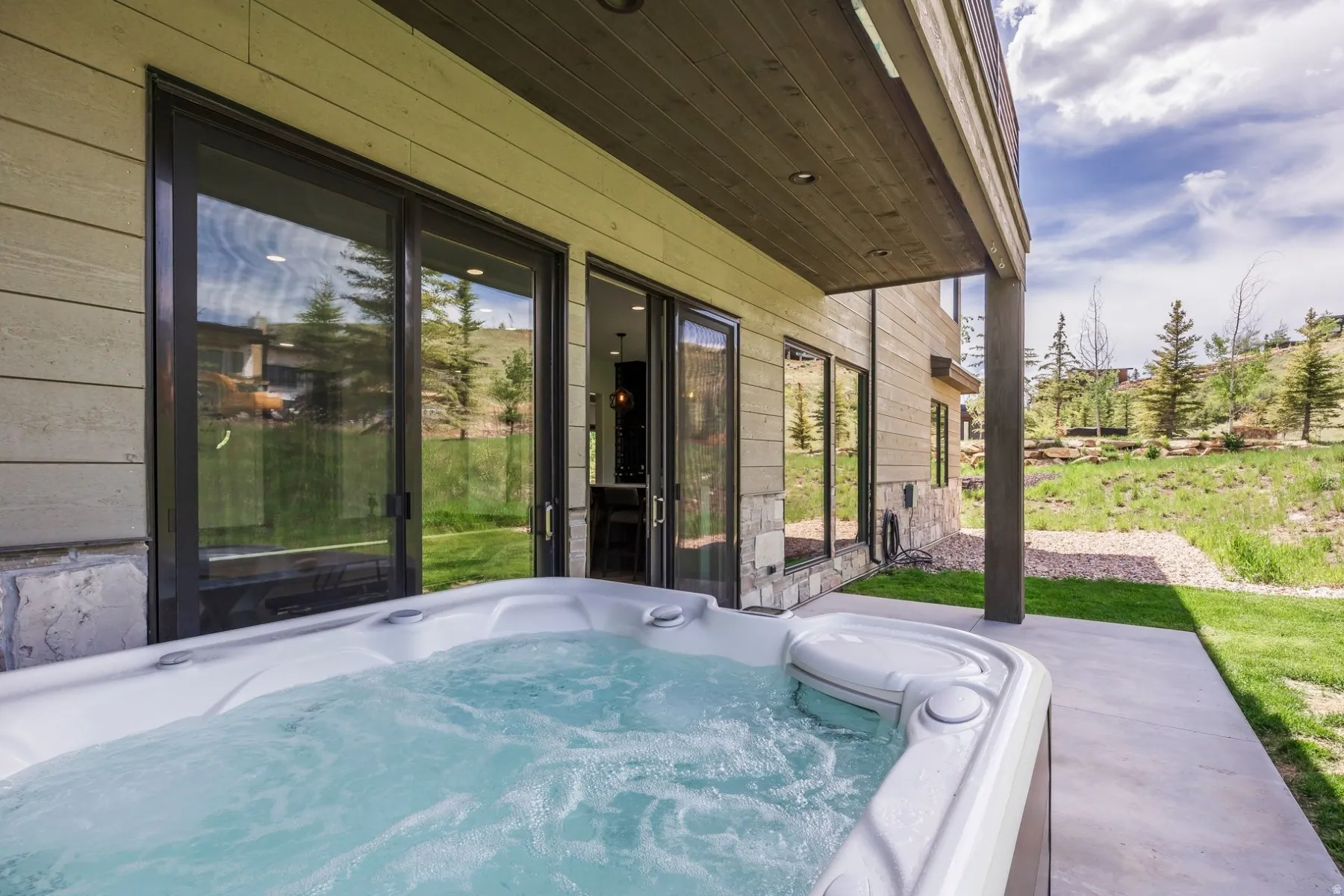 Outdoor hot tub on a concrete patio with sliding glass doors, surrounded by natural landscaping and stone accents