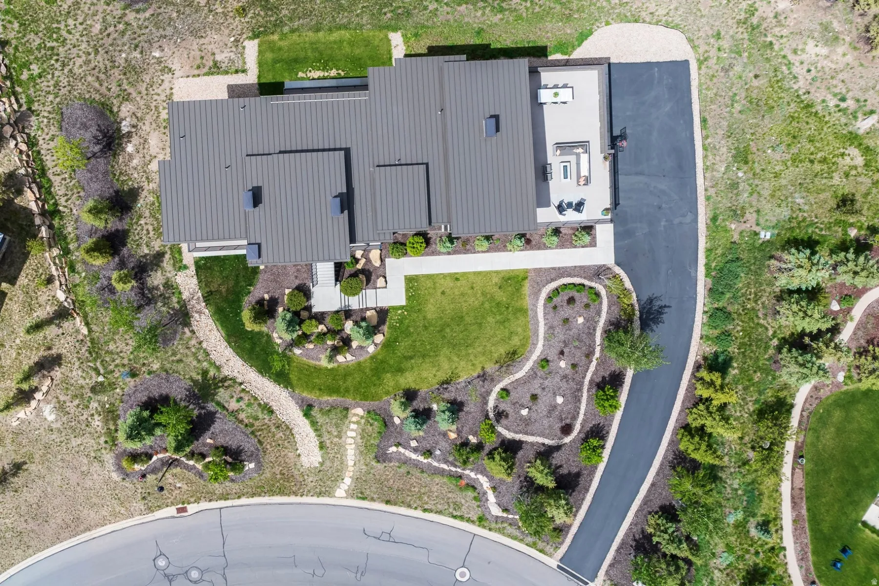Multi-level roofline with metal roofing, extensive hardscaping, a concrete patio, a paved driveway, and varied landscaping