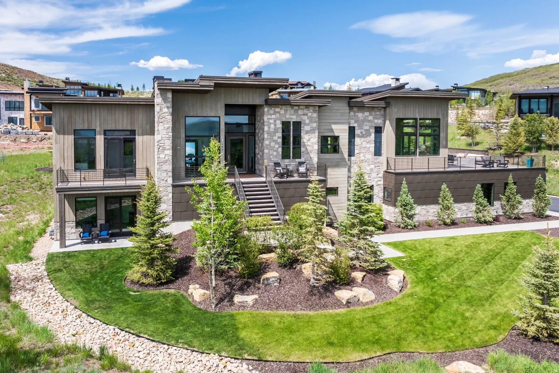 Contemporary architectural design featuring stone and wood-look siding, multiple outdoor balconies, a large ground-level patio, and a landscaped yard with mature plantings