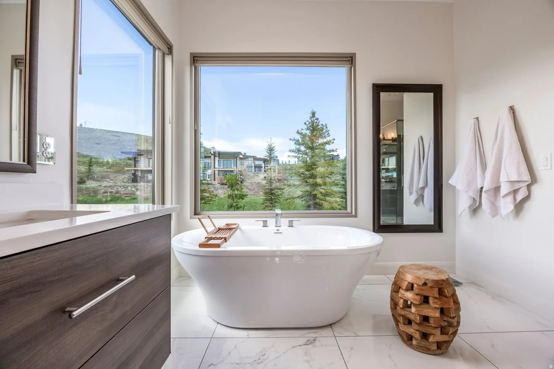 Freestanding soaking tub, wood-finish vanity with countertop, large format floor tiling, expansive windows, and a full-length wall mirror