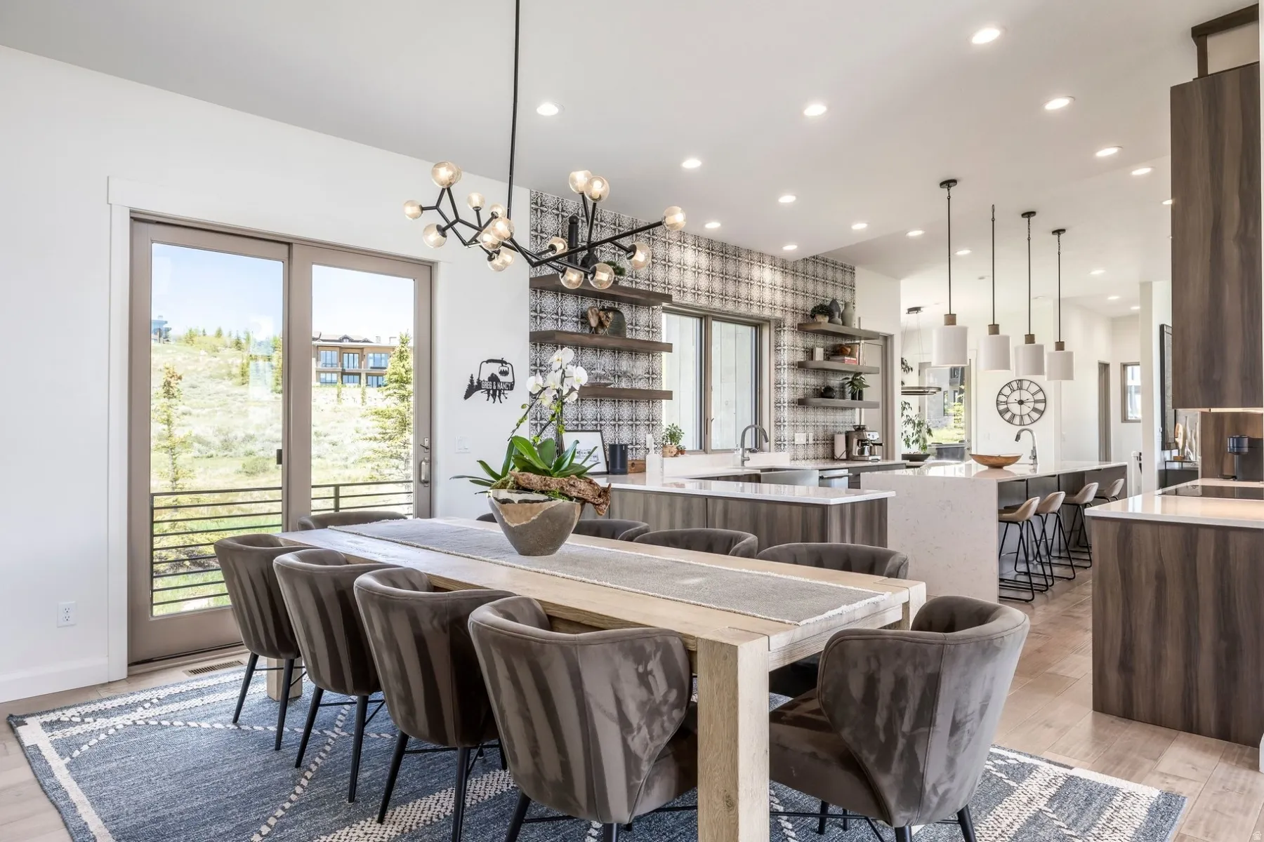 Modern open-concept living area with a contemporary chandelier, sliding glass door leading to an exterior balcony, light wood-finish flooring, and a kitchen featuring a patterned tile backsplash and white countertops