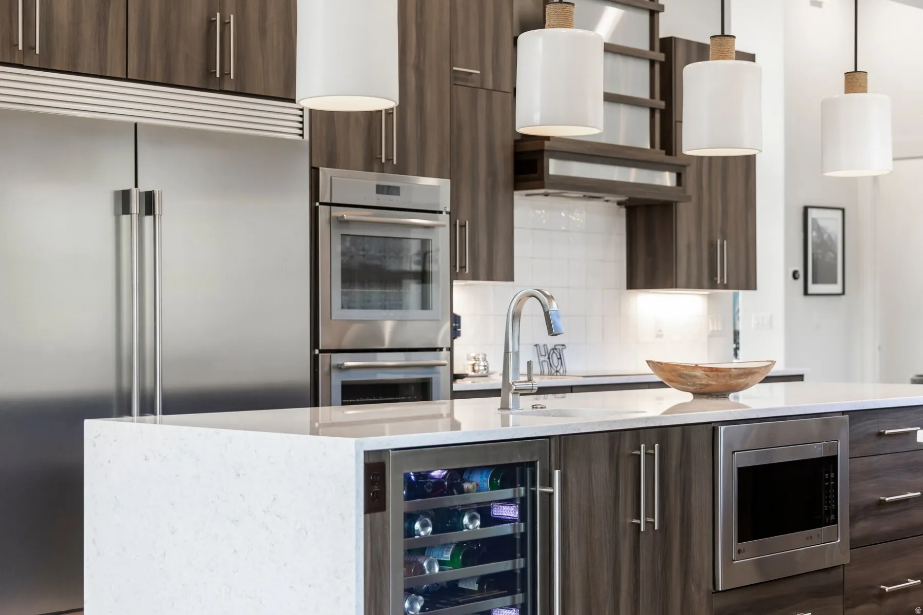 Kitchen featuring stainless steel appliances, wood-finish cabinetry, a white subway tile backsplash, a kitchen island with a built-in wine refrigerator, and pendant lighting