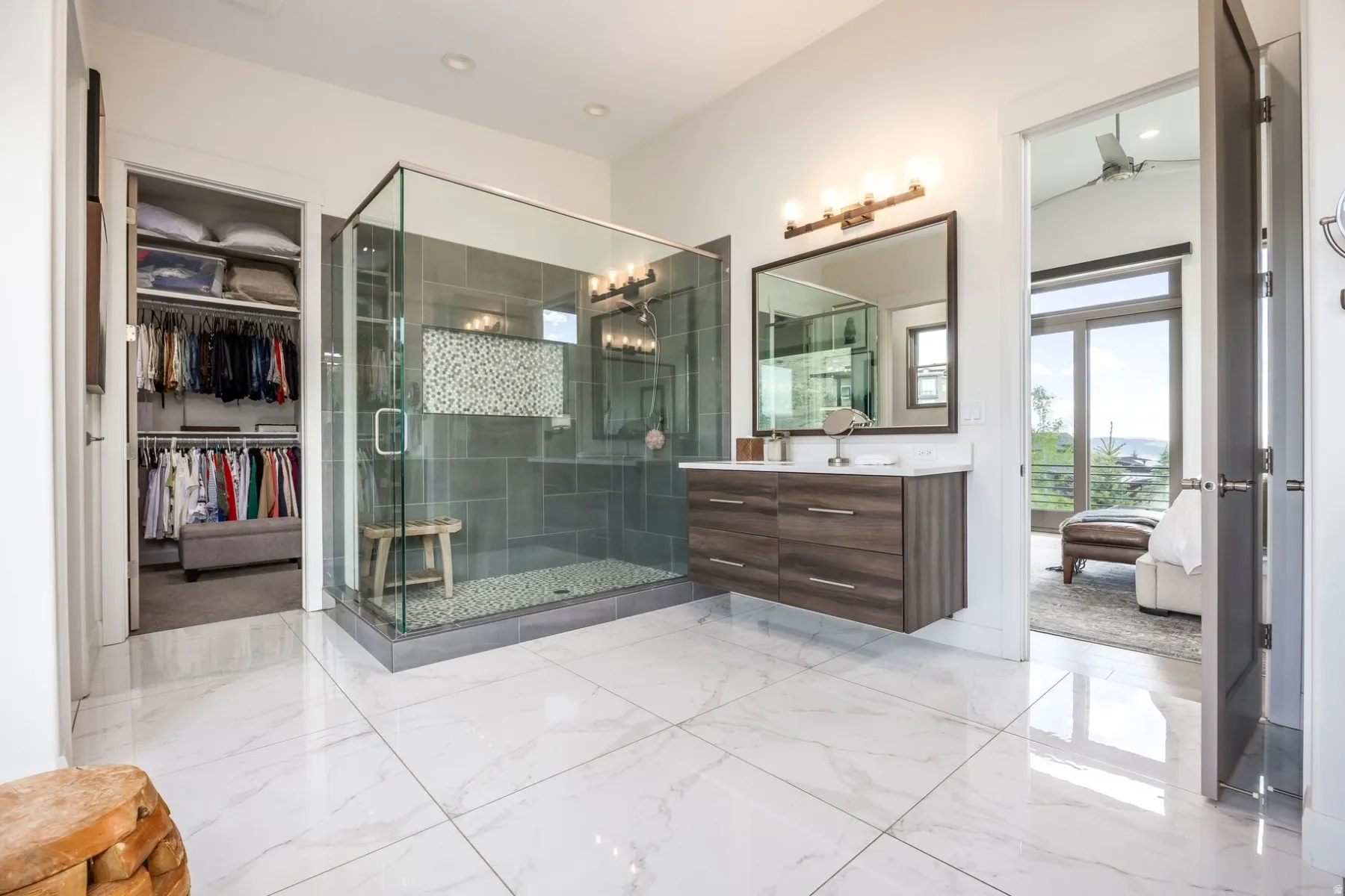 White tile flooring, glass-enclosed shower with pebble floor, floating wood-finish vanity with countertop, and an adjacent walk-in closet