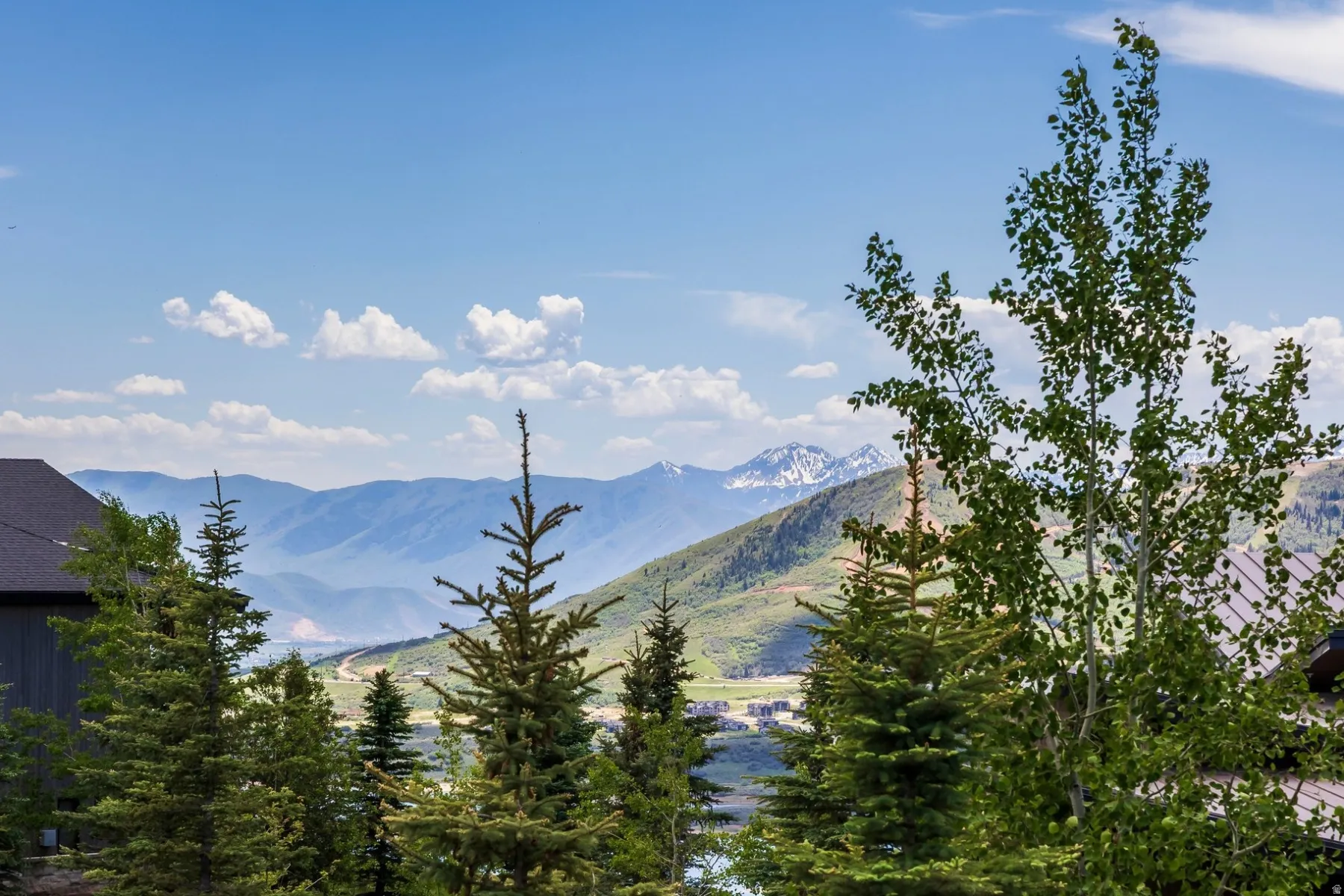 Mountainous backdrop with snow-capped peaks, surrounding evergreen trees, and partial residential structures