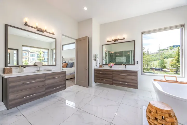 Dual floating wood-finish vanities with rectangular sinks, a freestanding bathtub, large format tile flooring, and a broad window