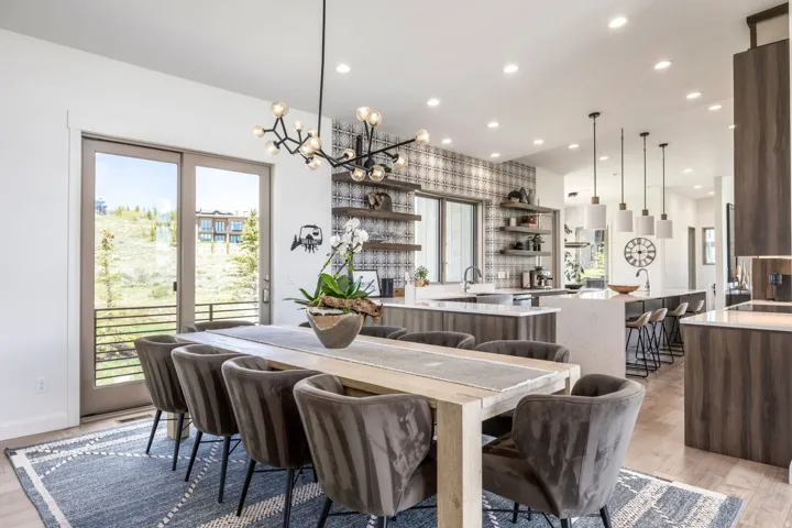 Modern open-concept living area with a contemporary chandelier, sliding glass door leading to an exterior balcony, light wood-finish flooring, and a kitchen featuring a patterned tile backsplash and white countertops