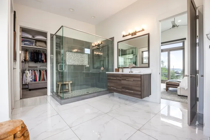 White tile flooring, glass-enclosed shower with pebble floor, floating wood-finish vanity with countertop, and an adjacent walk-in closet