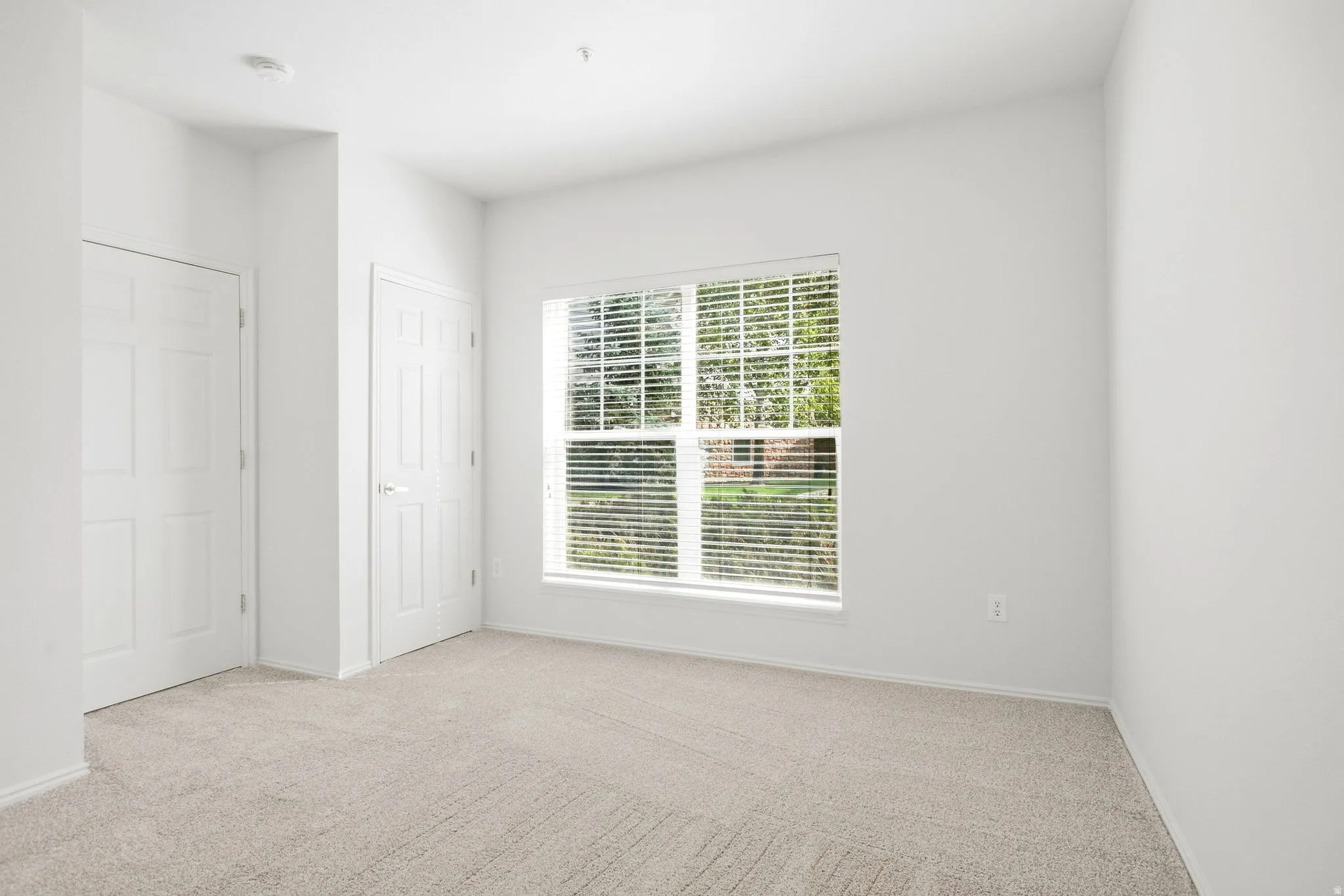 Neutral tone carpeting, a large window with blinds, and two paneled doors with chrome hardware