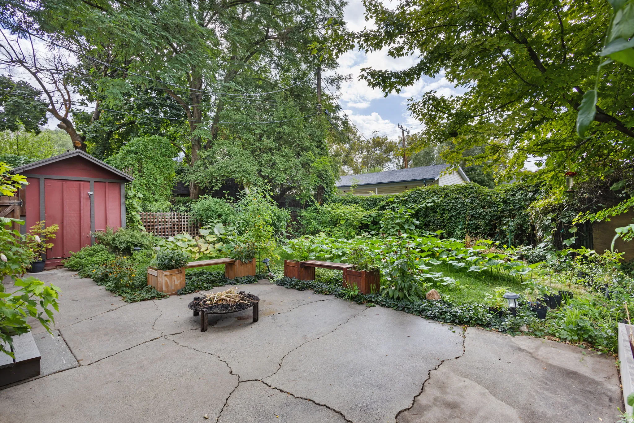 Expansive backyard featuring a concrete patio, a red storage shed, mature trees, built in benches, and a fire pit.