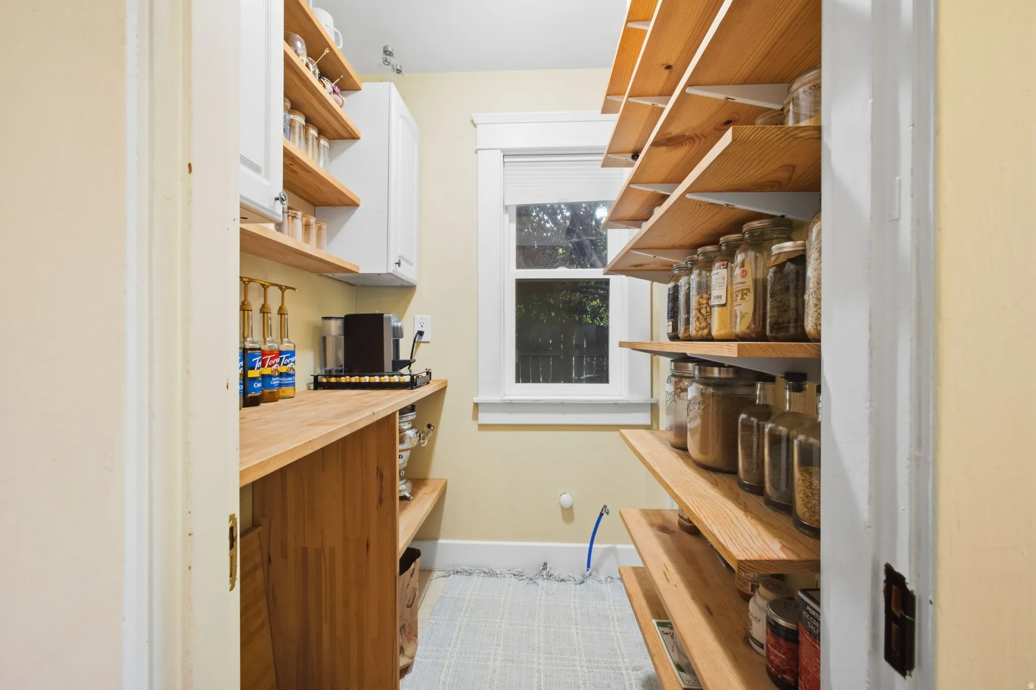 Butlers pantry includes built-in wood shelving, window with natural light, wood countertop, overhead cabinetry, water and drain hookups for an added sink, and light-colored flooring.