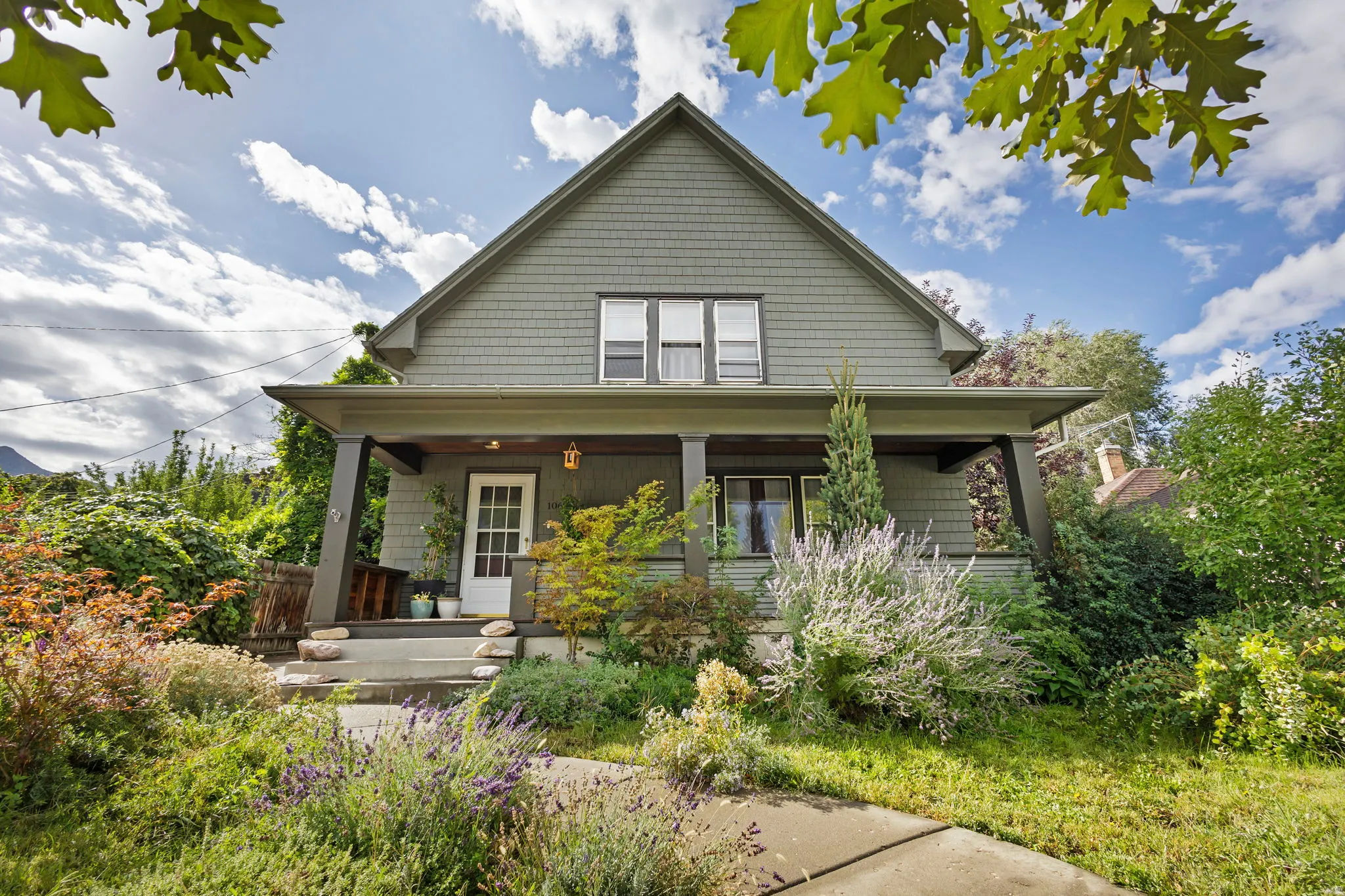 Gabled roofline, shake shingle siding, covered front porch with pillars, mature landscaping, and a concrete pathway.