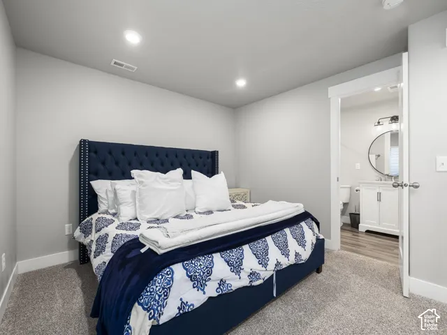 Carpeted bedroom featuring recessed lighting and a doorway leading to a bathroom with wood-finish flooring, a white vanity, and a circular mirror