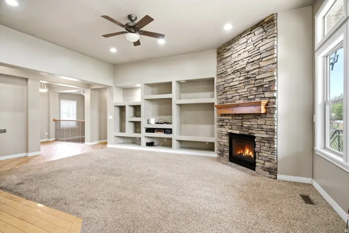 Stone-clad fireplace with a wood mantel, built-in shelving, ceiling fan, recessed lighting, and large windows