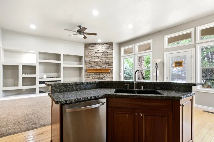 Kitchen island with stone countertop and integrated sink, stainless steel dishwasher, wood-finish flooring, a stacked stone fireplace with wood mantel, and built-in shelving